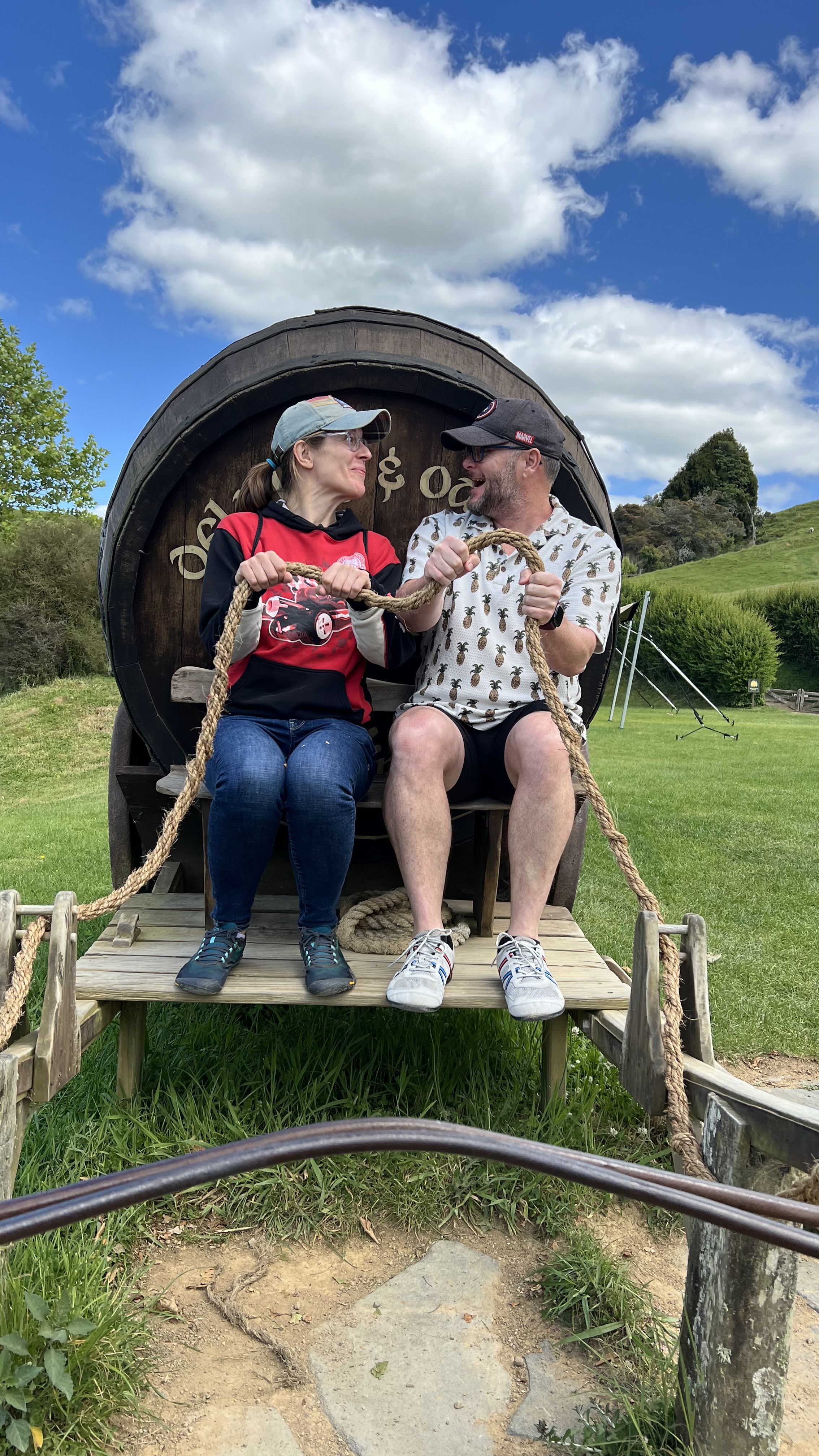 The Donegans at Hobbiton New Zealand adventure photo for Financial Independence Alan Donegan and Katie Donegan visiting Hobbiton in New Zealand riding a wooden horse cart beside a hobbit hole.