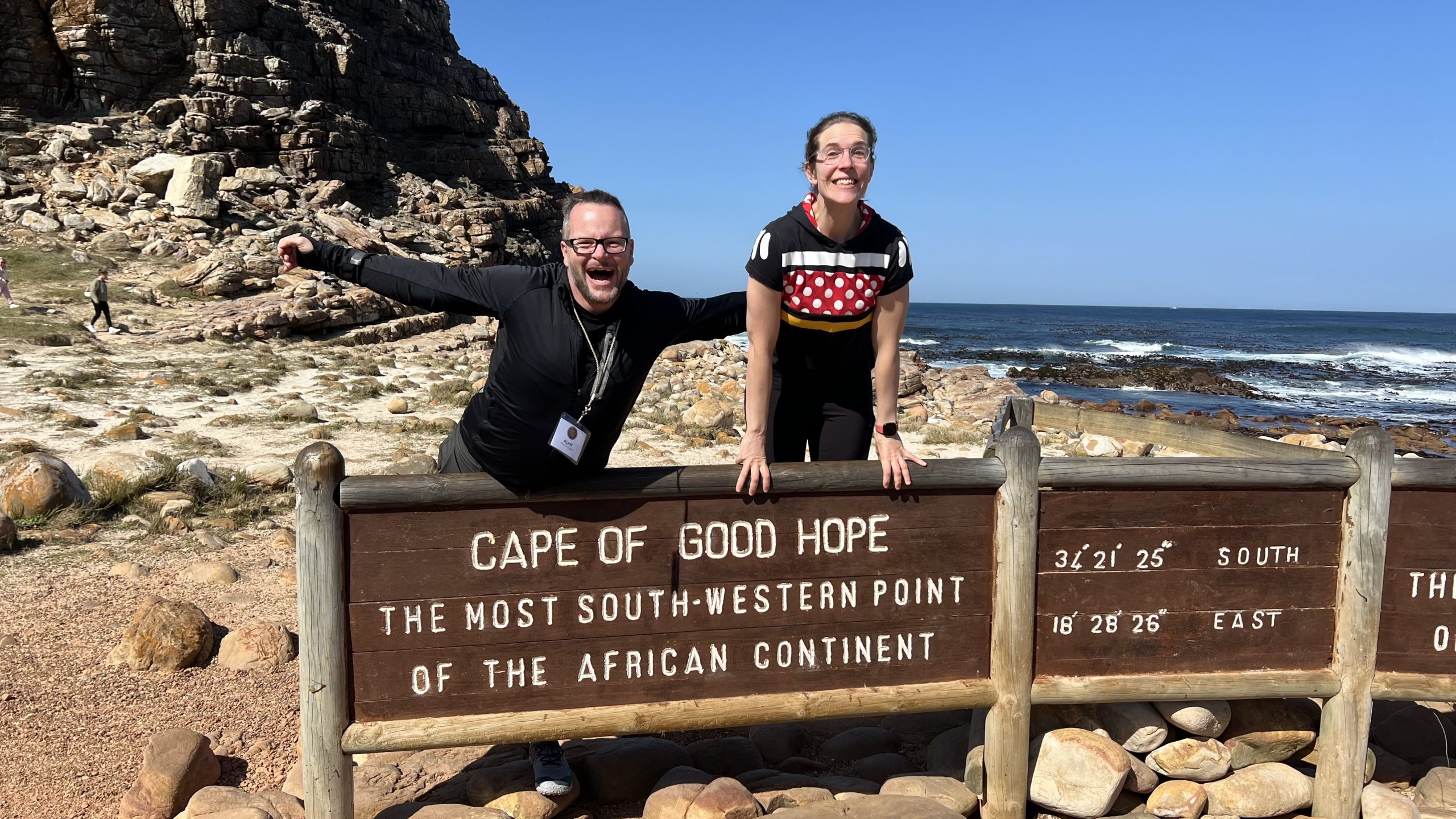 Standing at the Cape of Good Hope sign, the most south-western point of the African continent, with rocky coastline and ocean backdrop.